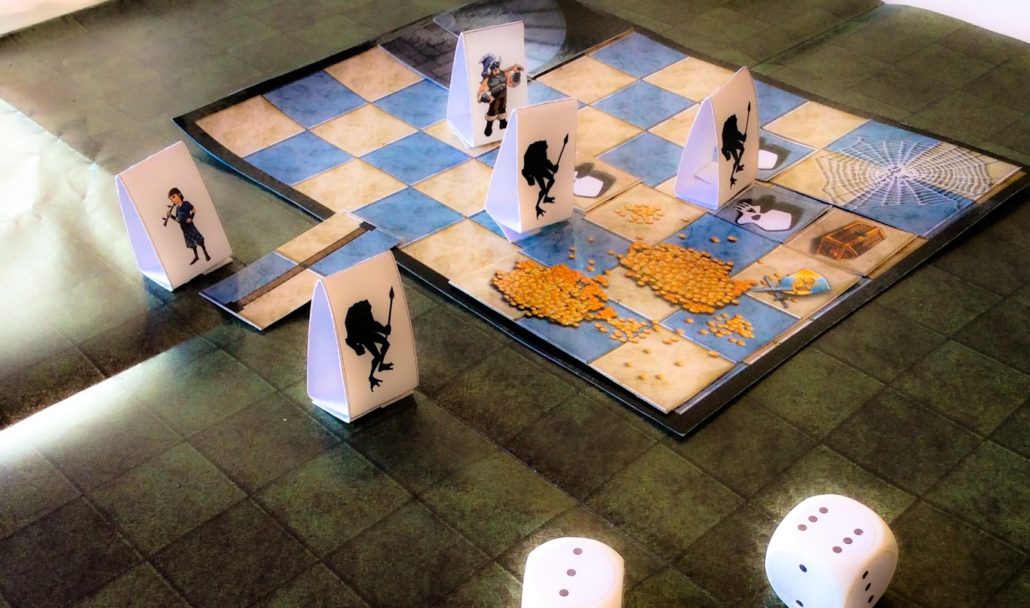 A imagem mostra o jogo de tabuleiro RPG Quest, um RPG para crianças. Há um tabuleiro quadrado com peças de papel dobradas, representando personagens e monstros. No canto inferior direito, há dois dados brancos de seis lados. O cenário do tabuleiro é uma masmorra com quadrados azuis e azulejos cinzentos, e algumas partes mostram teias de aranha e tesouros.