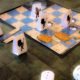 A imagem mostra o jogo de tabuleiro RPG Quest, um RPG para crianças. Há um tabuleiro quadrado com peças de papel dobradas, representando personagens e monstros. No canto inferior direito, há dois dados brancos de seis lados. O cenário do tabuleiro é uma masmorra com quadrados azuis e azulejos cinzentos, e algumas partes mostram teias de aranha e tesouros.