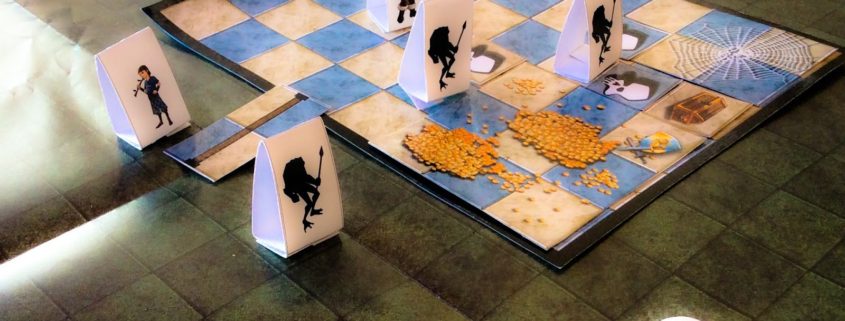 A imagem mostra o jogo de tabuleiro RPG Quest, um RPG para crianças. Há um tabuleiro quadrado com peças de papel dobradas, representando personagens e monstros. No canto inferior direito, há dois dados brancos de seis lados. O cenário do tabuleiro é uma masmorra com quadrados azuis e azulejos cinzentos, e algumas partes mostram teias de aranha e tesouros.