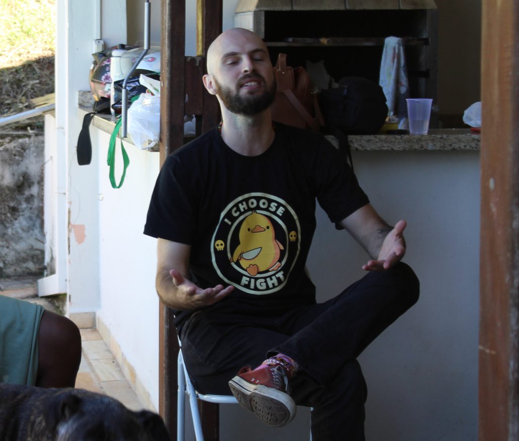Pessoa careca e com barba, sentada em uma cadeira de plástico branco, de olhos fechados e mãos abertas em um gesto expressivo, como se estivesse falando ou refletindo intensamente. Usa camiseta preta com a estampa de um pato amarelo e a frase “I CHOOSE FIGHT”. Está em uma área externa com churrasqueira e objetos ao fundo, transmitindo um clima descontraído e informal.