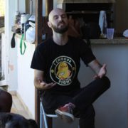 Respira fundo Pessoa careca e com barba, sentada em uma cadeira de plástico branco, de olhos fechados e mãos abertas em um gesto expressivo, como se estivesse falando ou refletindo intensamente. Usa camiseta preta com a estampa de um pato amarelo e a frase “I CHOOSE FIGHT”. Está em uma área externa com churrasqueira e objetos ao fundo, transmitindo um clima descontraído e informal.