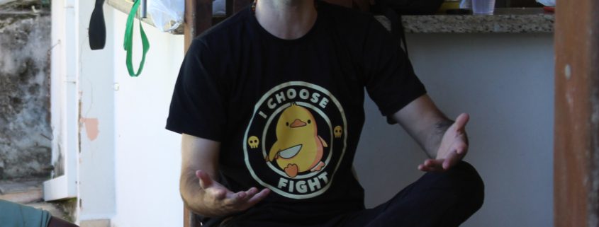 Pessoa careca e com barba, sentada em uma cadeira de plástico branco, de olhos fechados e mãos abertas em um gesto expressivo, como se estivesse falando ou refletindo intensamente. Usa camiseta preta com a estampa de um pato amarelo e a frase “I CHOOSE FIGHT”. Está em uma área externa com churrasqueira e objetos ao fundo, transmitindo um clima descontraído e informal.