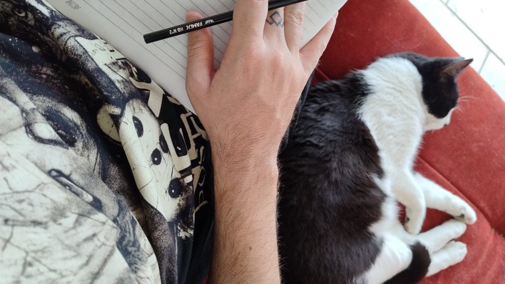 Foto de uma mão segurando lápis, com uma folha embaixo, e um gato deitado ao lado da pessoa que está escrevendo.