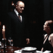 Cena do filme Hannibal (2001). Um homem de terno escuro (Dr. Hannibal Lecter, interpretado por Anthony Hopkins) está em pé ao lado de uma mesa de jantar elegantemente posta, com taças de vinho e velas acesas. Ele encara uma mulher (Clarice Starling, interpretada por Julianne Moore), que está sentada à mesa usando um vestido preto. A iluminação é baixa e dramática, criando um clima tenso e sombrio. A expressão dos personagens sugere uma conversa séria ou intimidadora.