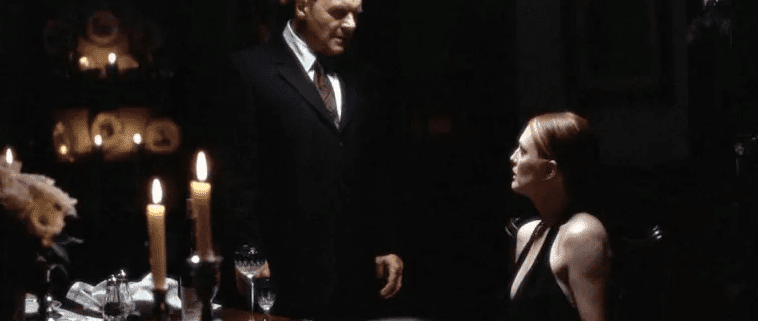 Cena do filme Hannibal (2001). Um homem de terno escuro (Dr. Hannibal Lecter, interpretado por Anthony Hopkins) está em pé ao lado de uma mesa de jantar elegantemente posta, com taças de vinho e velas acesas. Ele encara uma mulher (Clarice Starling, interpretada por Julianne Moore), que está sentada à mesa usando um vestido preto. A iluminação é baixa e dramática, criando um clima tenso e sombrio. A expressão dos personagens sugere uma conversa séria ou intimidadora.