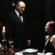 Cena do filme Hannibal (2001). Um homem de terno escuro (Dr. Hannibal Lecter, interpretado por Anthony Hopkins) está em pé ao lado de uma mesa de jantar elegantemente posta, com taças de vinho e velas acesas. Ele encara uma mulher (Clarice Starling, interpretada por Julianne Moore), que está sentada à mesa usando um vestido preto. A iluminação é baixa e dramática, criando um clima tenso e sombrio. A expressão dos personagens sugere uma conversa séria ou intimidadora.