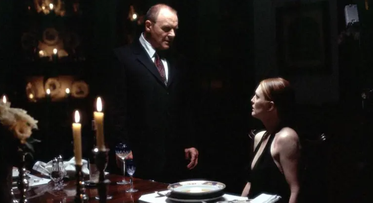 Cena do filme Hannibal (2001). Um homem de terno escuro (Dr. Hannibal Lecter, interpretado por Anthony Hopkins) está em pé ao lado de uma mesa de jantar elegantemente posta, com taças de vinho e velas acesas. Ele encara uma mulher (Clarice Starling, interpretada por Julianne Moore), que está sentada à mesa usando um vestido preto. A iluminação é baixa e dramática, criando um clima tenso e sombrio. A expressão dos personagens sugere uma conversa séria ou intimidadora.
