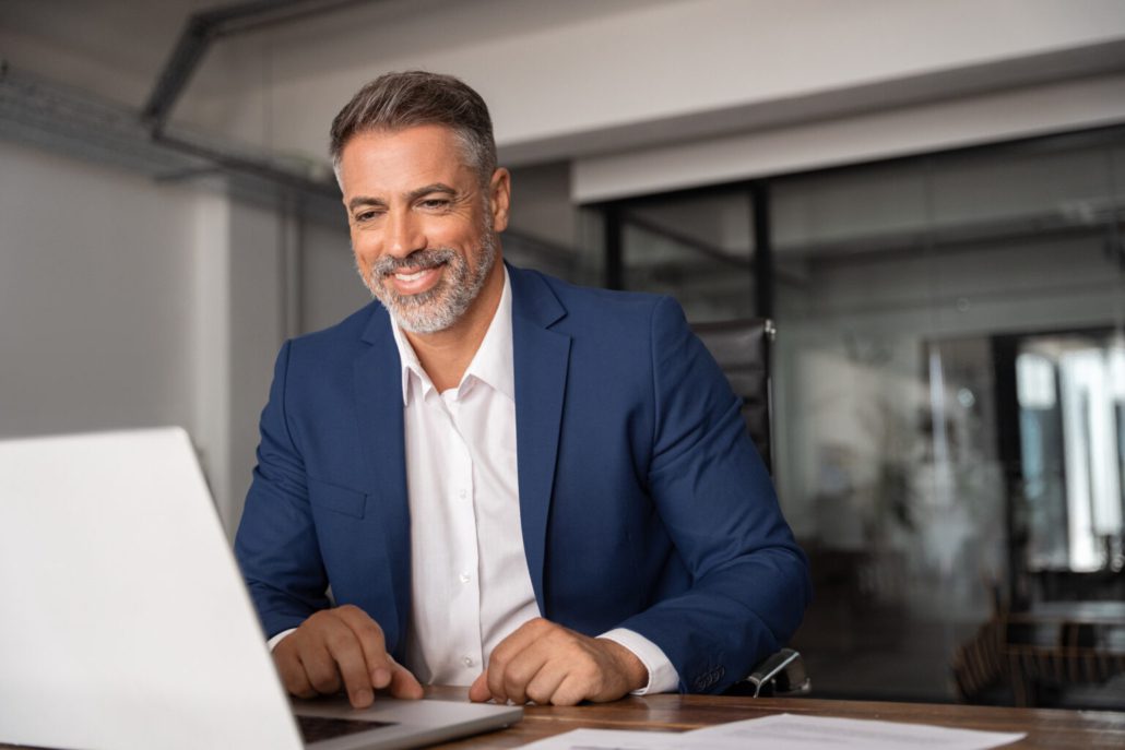 A imagem mostra um homem executivo sentado, de frente a uma mesa com notebook.