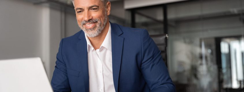A imagem mostra um homem executivo sentado, de frente a uma mesa com notebook.