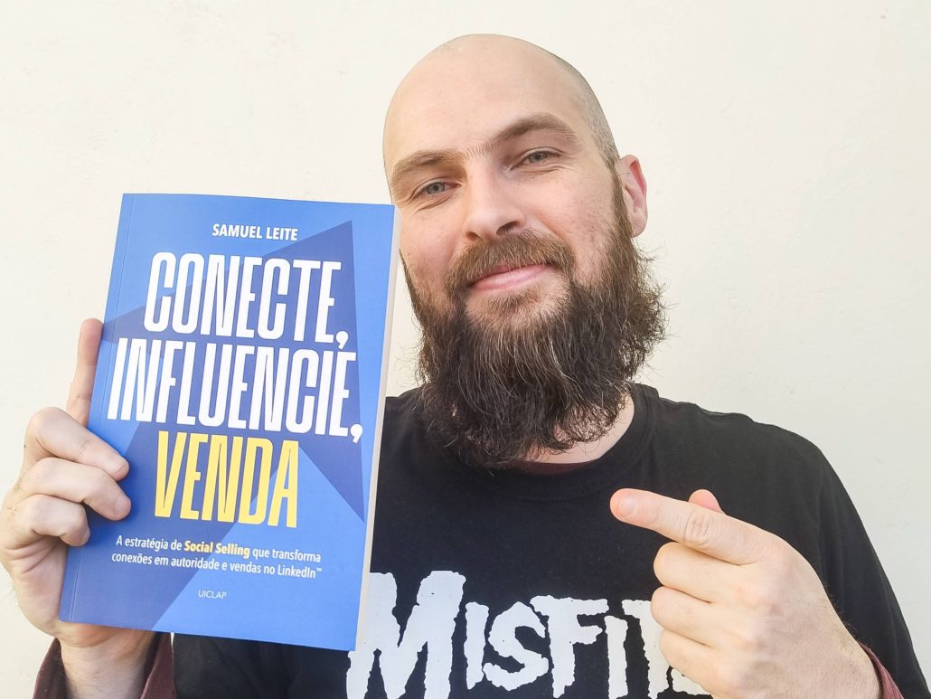 Homem branco, de barba cheia e cabeça raspada, sorri levemente e segura com uma mão o livro “Conecte, Influencie, Venda”, de Samuel Leite, enquanto aponta para ele com a outra mão. A capa do livro é azul com letras brancas e amarelas. Ele veste uma camiseta preta com estampa da banda Misfits. O fundo é neutro, de parede clara.