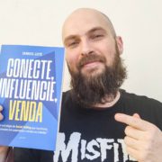 PSX_20250729_142738 Homem branco, de barba cheia e cabeça raspada, sorri levemente e segura com uma mão o livro “Conecte, Influencie, Venda”, de Samuel Leite, enquanto aponta para ele com a outra mão. A capa do livro é azul com letras brancas e amarelas. Ele veste uma camiseta preta com estampa da banda Misfits. O fundo é neutro, de parede clara.