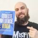 Homem branco, de barba cheia e cabeça raspada, sorri levemente e segura com uma mão o livro “Conecte, Influencie, Venda”, de Samuel Leite, enquanto aponta para ele com a outra mão. A capa do livro é azul com letras brancas e amarelas. Ele veste uma camiseta preta com estampa da banda Misfits. O fundo é neutro, de parede clara.