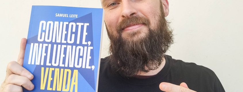 Homem branco, de barba cheia e cabeça raspada, sorri levemente e segura com uma mão o livro “Conecte, Influencie, Venda”, de Samuel Leite, enquanto aponta para ele com a outra mão. A capa do livro é azul com letras brancas e amarelas. Ele veste uma camiseta preta com estampa da banda Misfits. O fundo é neutro, de parede clara.