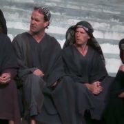 A imagem mostra uma cena do filme "A Vida de Brian". Quatro pessoas, três homens e uma mulher, estão sentadas em degraus de pedra. Todos estão vestidos com túnicas escuras e coberturas para a cabeça, com expressões pensativas.