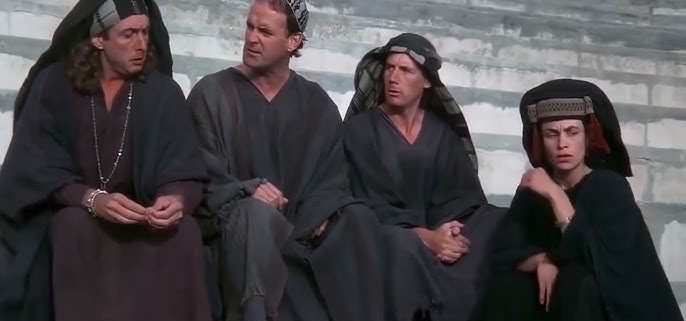 A imagem mostra uma cena do filme "A Vida de Brian". Quatro pessoas, três homens e uma mulher, estão sentadas em degraus de pedra. Todos estão vestidos com túnicas escuras e coberturas para a cabeça, com expressões pensativas.