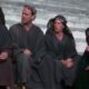 A imagem mostra uma cena do filme "A Vida de Brian". Quatro pessoas, três homens e uma mulher, estão sentadas em degraus de pedra. Todos estão vestidos com túnicas escuras e coberturas para a cabeça, com expressões pensativas.
