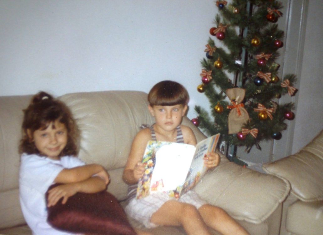 Na foto, duas crianças estão sentadas em um sofá: uma sorri abraçando uma almofada, minha prima, enquanto a outra segura o livro “A fada que tinha ideias” (eu). Ao lado delas há uma árvore de Natal decorada com bolas coloridas e laços, a foto é a cara dos anos 90.