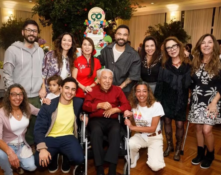 Foto do aniversário do Mauricio de Sousa, onde ele está em uma cadeira de rodas no centro da imagem cercado da família.