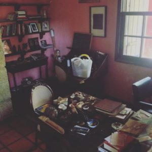 Escritório da escritora Hilda Hilst na Casa do Sol, em Campinas. O ambiente tem paredes em tom rosado, uma mesa cheia de papéis, livros e objetos pessoais, estantes com livros e uma janela por onde entra luz natural.