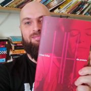 Homem de barba e cabeça raspada segura um livro de capa vermelha diante do rosto. Na capa, lê-se “hilda hilst” e “da poesia”. Ao fundo, há uma estante com muitos livros.