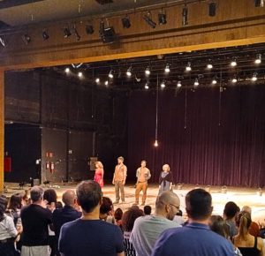 Foto interna do Teatro Castro Mendes, em Campinas, vista a partir da plateia. No palco, após a apresentação, estão quatro atores alinhados, da esquerda para a direita: Camila Nhary, Iano Salomão, Armando Babaioff e Denise Del Vecchio. Ao fundo, há uma cortina escura e um cenário simples. Em primeiro plano, o público aparece em pé, aplaudindo.