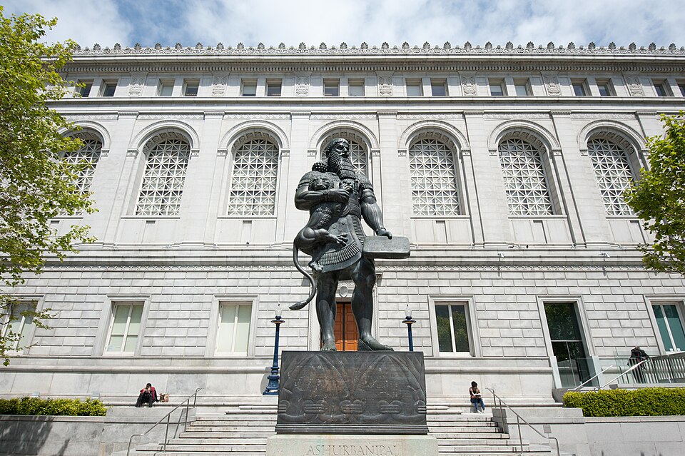 A imagem mostra a estátua de Assurbanípal em São Francisco. A escultura retrata um rei da antiga Mesopotâmia segurando um leão e um livro, sobre um pedestal em frente a um edifício.