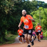 Homem careca com barba preta em destaque, correndo em uma estrada de terra em meio à natureza durante uma prova de trail run. Ele veste camiseta laranja do evento e short preto. Ele sorri e faz um gesto de paz com a mão para a câmera enquanto corre. Ao fundo, outros corredores também participam da prova, cercados por vegetação verde no entorno da Fazenda Rio Acima, em Pedreira.