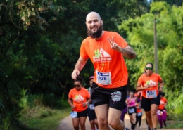 Homem careca com barba preta em destaque, correndo em uma estrada de terra em meio à natureza durante uma prova de trail run. Ele veste camiseta laranja do evento e short preto. Ele sorri e faz um gesto de paz com a mão para a câmera enquanto corre. Ao fundo, outros corredores também participam da prova, cercados por vegetação verde no entorno da Fazenda Rio Acima, em Pedreira.