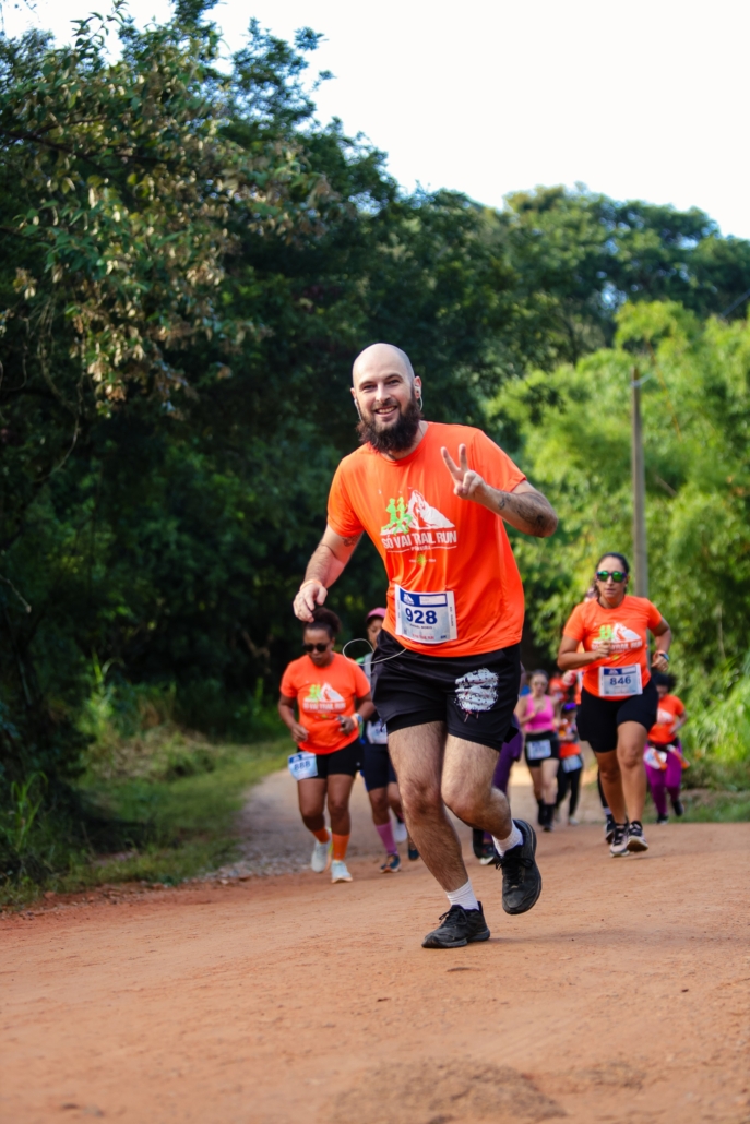 Homem careca com barba preta em destaque, correndo em uma estrada de terra em meio à natureza durante uma prova de trail run. Ele veste camiseta laranja do evento e short preto. Ele sorri e faz um gesto de paz com a mão para a câmera enquanto corre. Ao fundo, outros corredores também participam da prova, cercados por vegetação verde no entorno da Fazenda Rio Acima, em Pedreira.