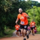 Homem careca com barba preta em destaque, correndo em uma estrada de terra em meio à natureza durante uma prova de trail run. Ele veste camiseta laranja do evento e short preto. Ele sorri e faz um gesto de paz com a mão para a câmera enquanto corre. Ao fundo, outros corredores também participam da prova, cercados por vegetação verde no entorno da Fazenda Rio Acima, em Pedreira.