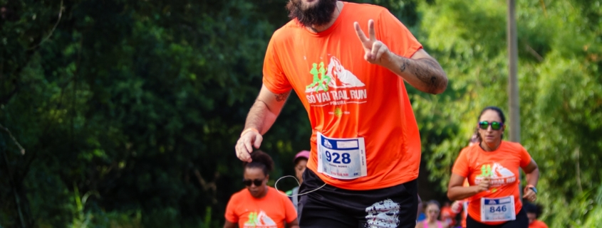 Homem careca com barba preta em destaque, correndo em uma estrada de terra em meio à natureza durante uma prova de trail run. Ele veste camiseta laranja do evento e short preto. Ele sorri e faz um gesto de paz com a mão para a câmera enquanto corre. Ao fundo, outros corredores também participam da prova, cercados por vegetação verde no entorno da Fazenda Rio Acima, em Pedreira.